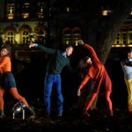 Flere personer, klædt i farverigt tøj, står i en bypark om aftenen. De er fotograferet midt i en dans, hvor de hver især strækker sig i akrobatiske stillinger