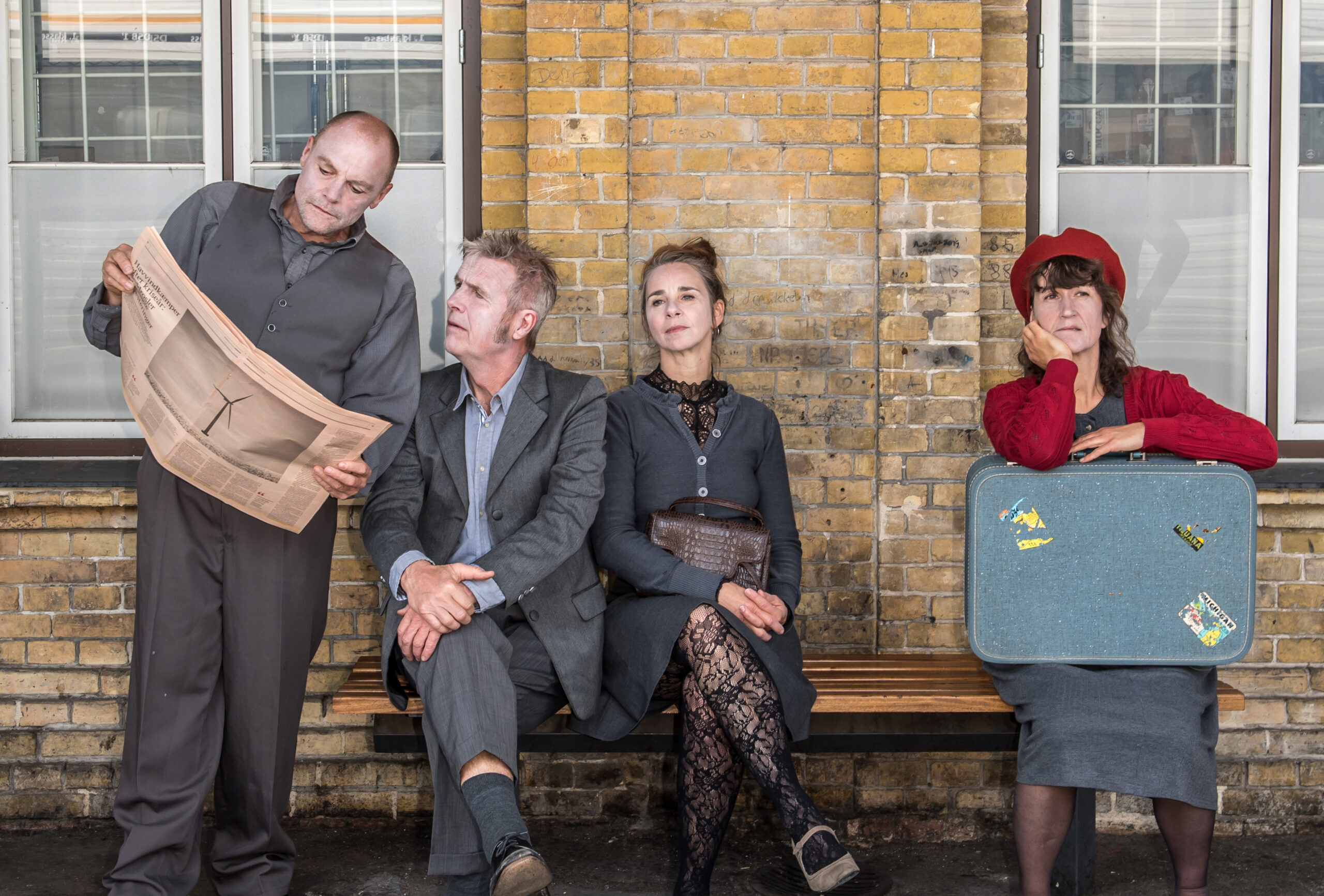 En mand står og læser avis. En mand sidder ved siden af ham på en bænk og læser med i avisen. To kvinder sidder også på bænken, og stirrer ud i luften. De ser ud til at være på en togstation.