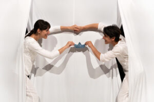 To kvinder holder en den samme papirbåd. De kigger frem bag et hvidt gardin og holder hver papirbåden i én hånd.