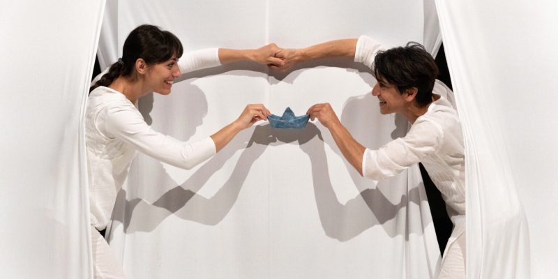 To kvinder holder en den samme papirbåd. De kigger frem bag et hvidt gardin og holder hver papirbåden i én hånd.